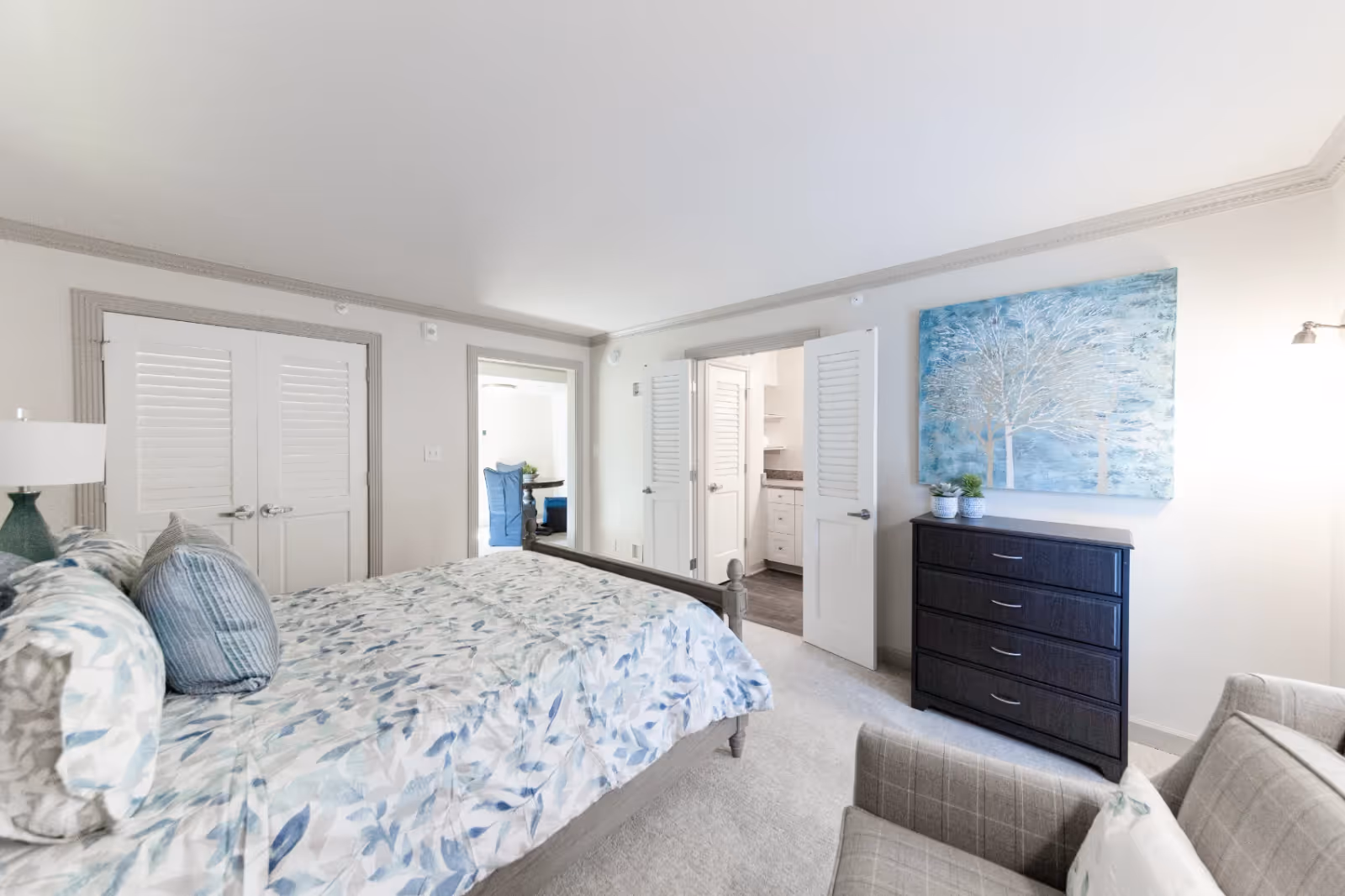A bright and airy bedroom with a bed covered in a floral patterned comforter and pillows. There is a dark wooden dresser with two small potted plants on top and a large blue and white painting of trees hanging above it. A gray armchair with a pillow is positioned in the corner. The room has white walls with gray trim, a double-door closet, and an open doorway leading to another room with a blue chair and a table.