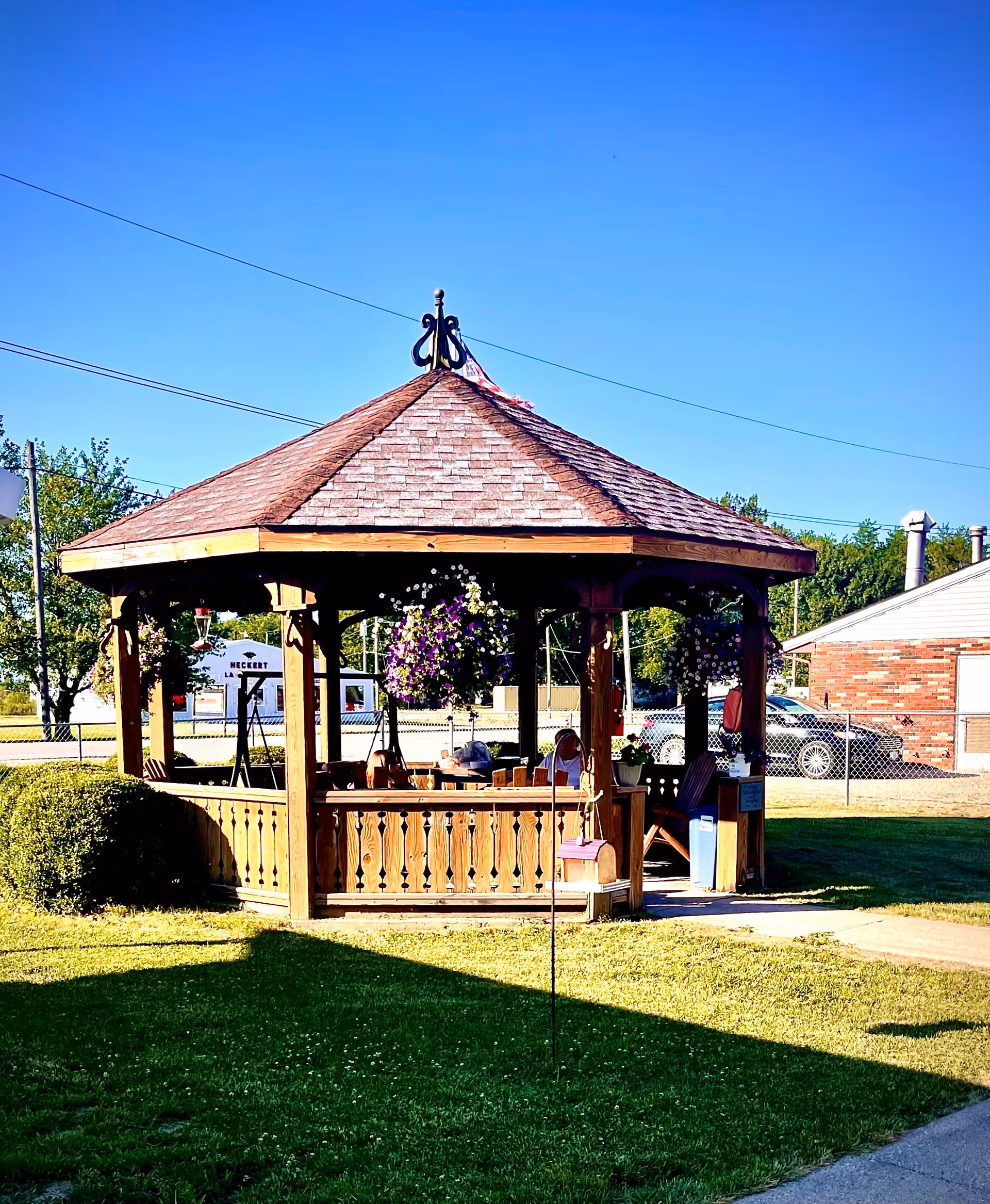 A wooden gazebo with a shingled roof and decorative finial on top, situated on a grassy area with hanging flower baskets. In the background, there is a chain-link fence, a brick building, and a clear blue sky.