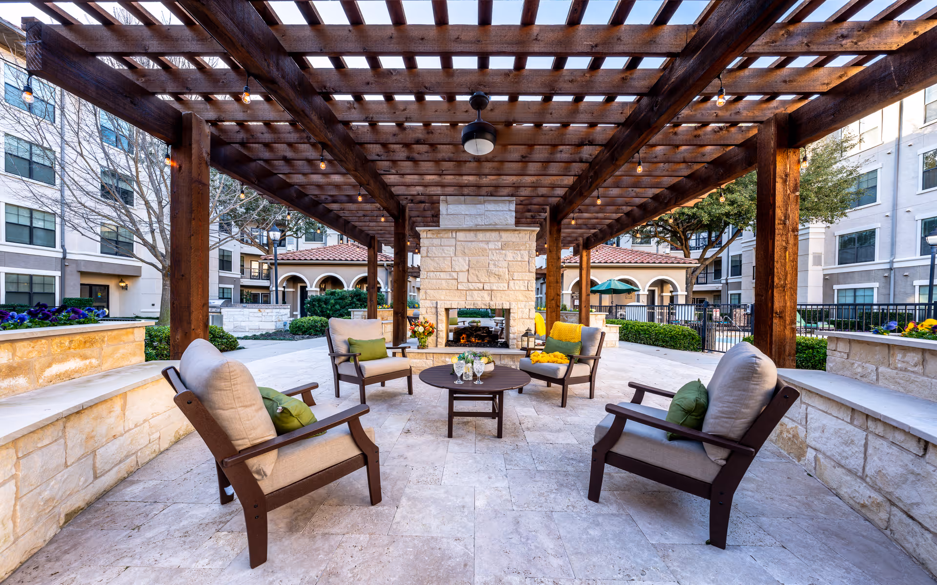 Outdoor seating area at Conservatory At Plano featuring four cushioned chairs arranged around a round table with drinks and flowers, under a wooden pergola with string lights. A stone fireplace is centered at the back, surrounded by landscaped greenery and multi-story residential buildings.