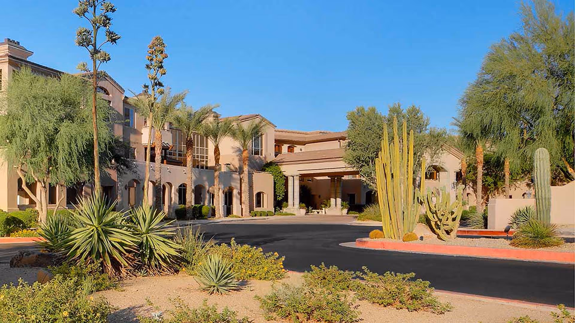 Exterior view of a senior living facility with desert landscaping including various cacti and palm trees under a clear blue sky.