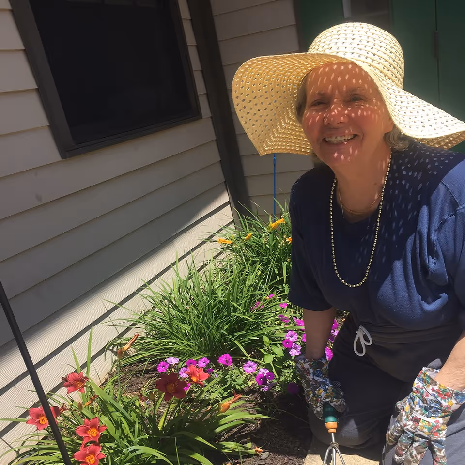 An elderly woman wearing a large straw hat, floral gardening gloves, and a dark blue shirt is smiling while gardening next to a house. She is kneeling beside a flower bed with blooming red, purple, and yellow flowers.