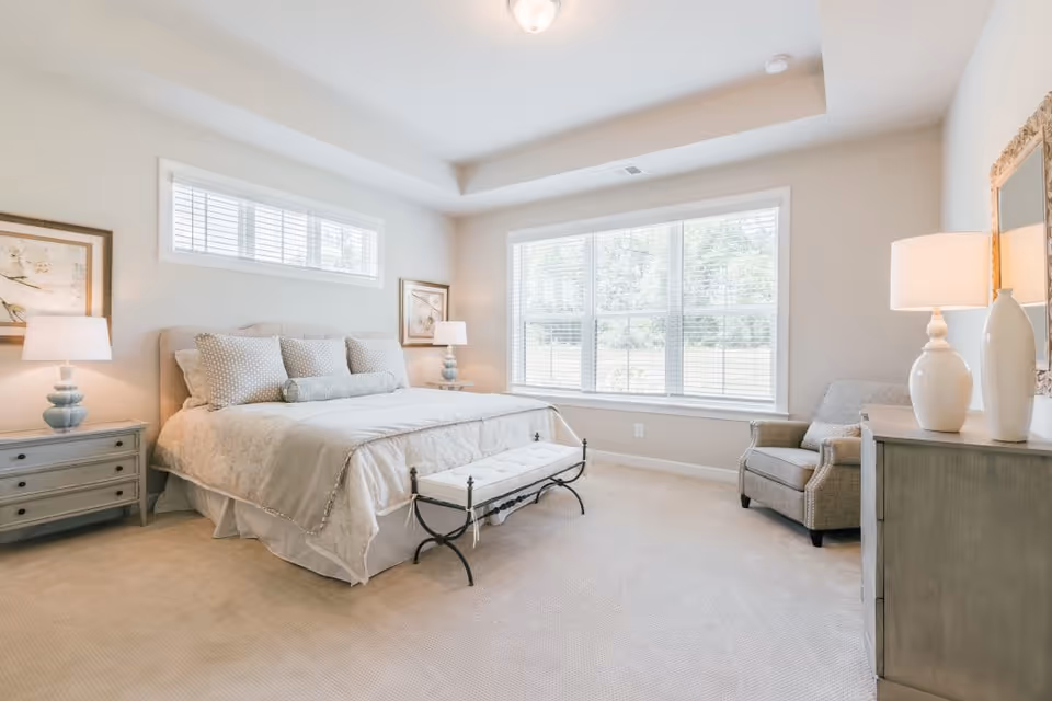 Bright, neutral-toned bedroom with a large bed, nightstands and lamps, a dresser and an armchair by a large window.