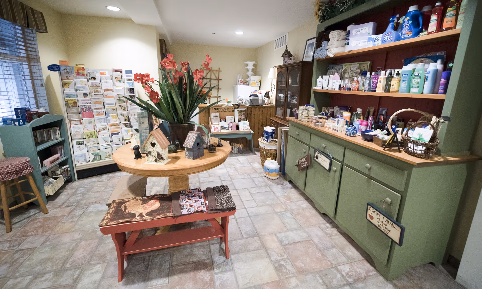 Interior view of a small shop or gift area in a senior living facility with a round wooden table in the center displaying decorative birdhouses and a vase with red flowers. To the right, there is a green cabinet filled with various personal care products and household items. In the background, there is a rack with greeting cards and other small items. The floor is tiled and the lighting is bright.