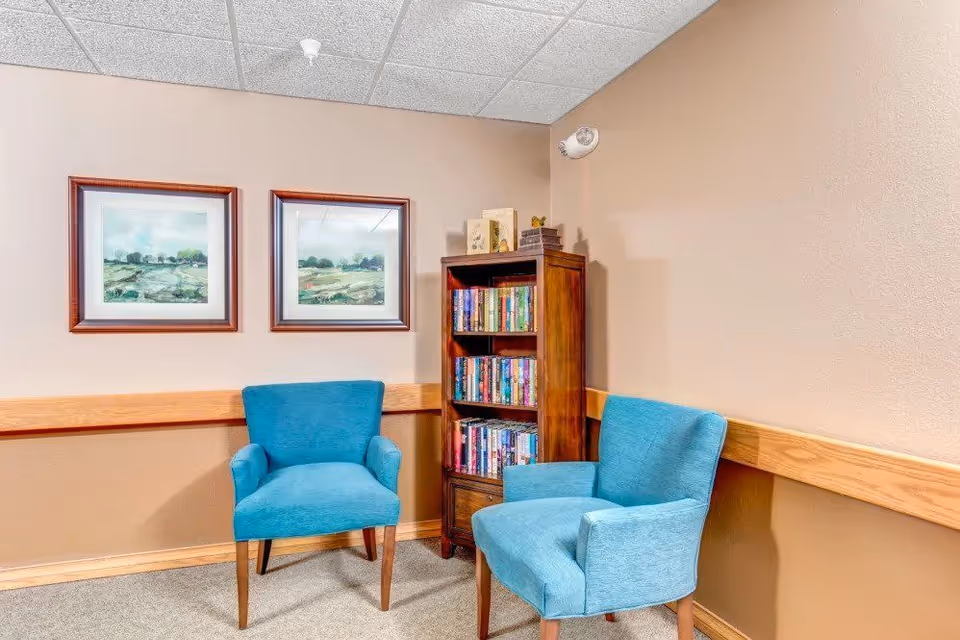 A cozy corner in a senior living facility with two blue upholstered armchairs facing each other. Between them is a wooden bookshelf filled with books. The walls are beige with a wooden chair rail, and two framed landscape paintings hang above one of the chairs.