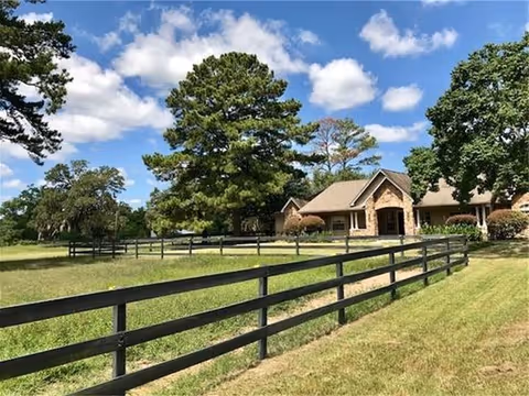 A single-story brick building with a gabled roof surrounded by trees and a large grassy area enclosed by a black wooden fence under a blue sky with scattered clouds.