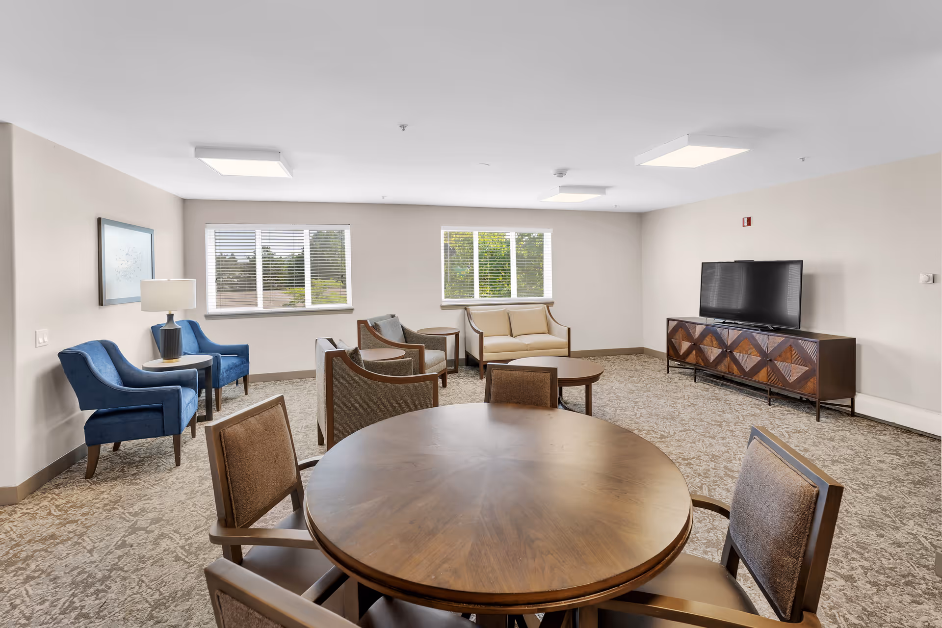 A spacious living room with a round wooden table and four chairs in the foreground. The room features two blue armchairs with a small round table and lamp between them on the left side. In the background, there are two beige armchairs with a small round table between them, a beige loveseat, and a large flat-screen TV on a wooden cabinet with a geometric pattern. The room has two large windows with white blinds letting in natural light, beige walls, and carpeted flooring.