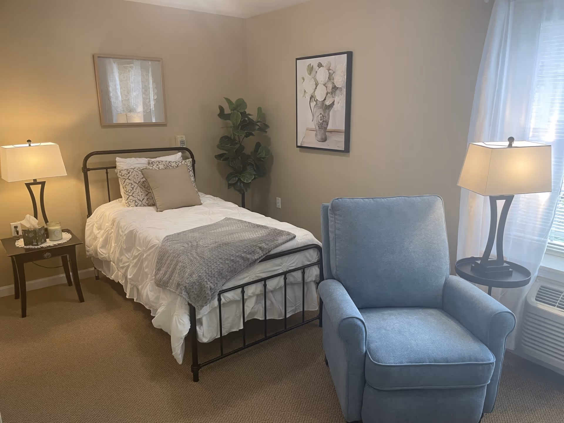 A cozy bedroom with a single bed featuring white bedding, decorative pillows, and a gray throw blanket. There is a blue upholstered armchair near a window with sheer white curtains. Two lamps on dark wooden side tables provide warm lighting. A framed floral painting hangs on the wall above the bed, and a green potted plant is placed in the corner.