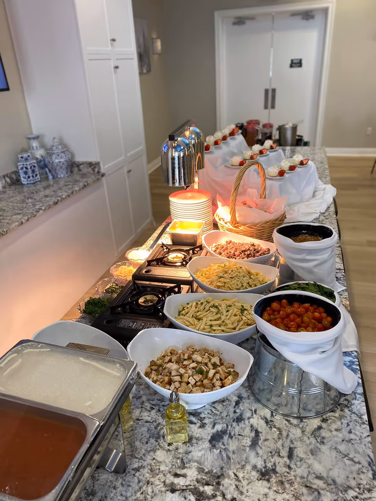 A buffet-style food setup on a granite countertop featuring bowls of pasta, diced chicken, cherry tomatoes, chopped meat, and various garnishes. There are plates stacked under a heat lamp, a basket with bread, and small dessert cups with whipped cream and strawberries arranged on a tiered display in the background. The setting appears to be indoors with white cabinets and double doors visible.