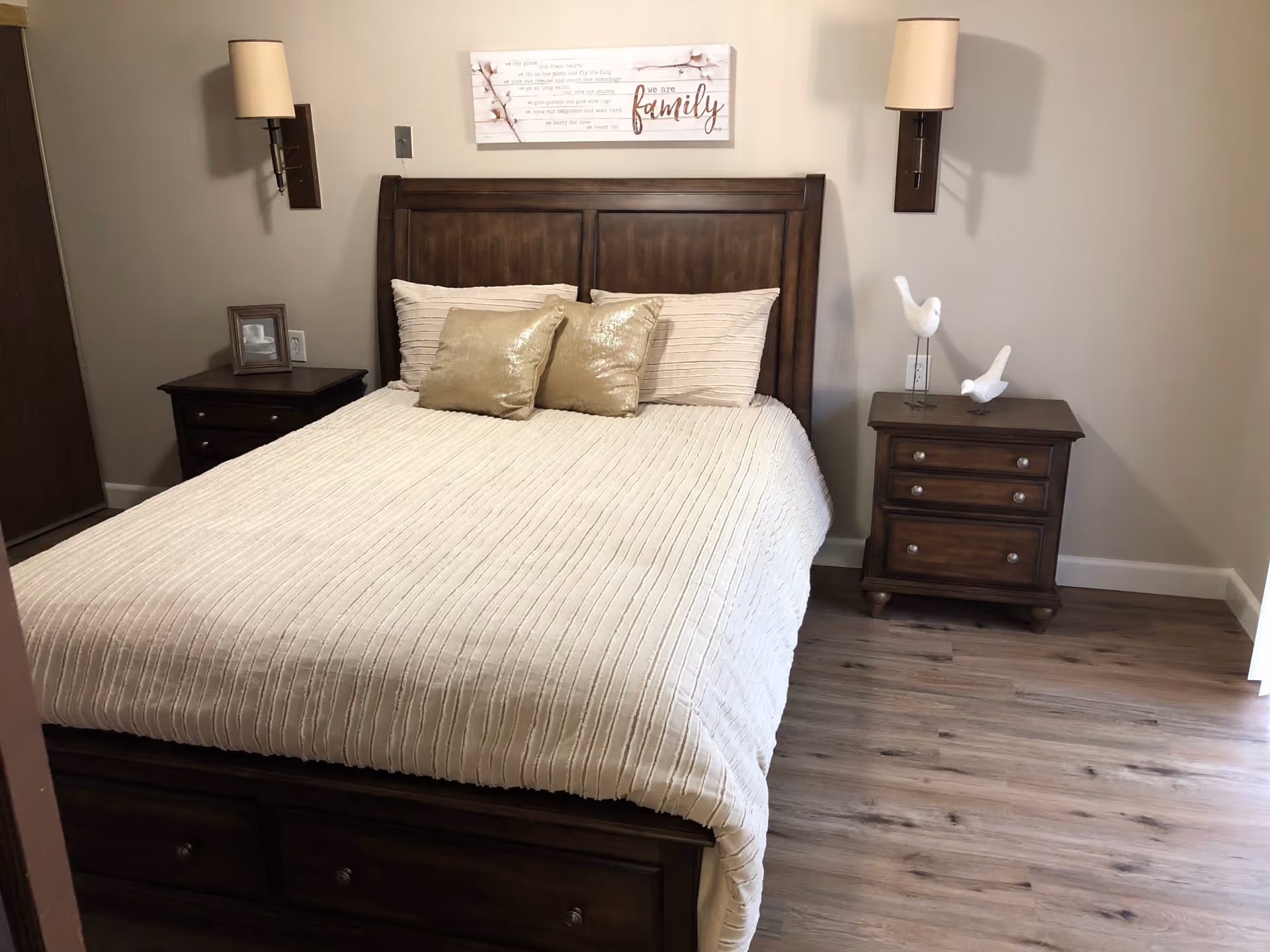 A neatly made bed with a wooden headboard and two gold pillows in a bedroom. There are two matching wooden nightstands on either side of the bed, each with a lamp mounted on the wall above. One nightstand has a framed photo, and the other has two decorative white bird figurines. Above the bed is a wall decoration with the word 'family' and a quote. The room has wood flooring and beige walls.