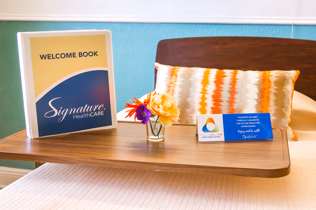 A wooden tray table on a bed with a decorative pillow in the background. On the tray is a welcome book with the Signature HealthCARE logo, a small glass vase with colorful flowers, and a card indicating the room has been carefully cleaned.