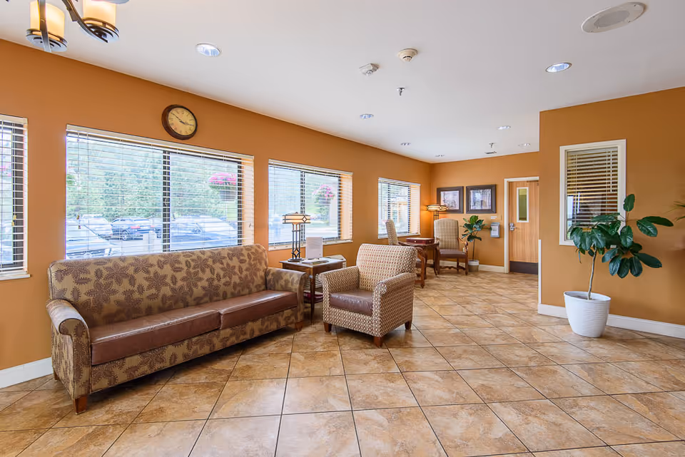A bright and spacious common area with tan walls and tiled floor. The room features a patterned sofa and armchair, a small table with a lamp, and two chairs with a table near the back wall. Large windows with blinds let in natural light, and there are potted plants and framed pictures on the walls.