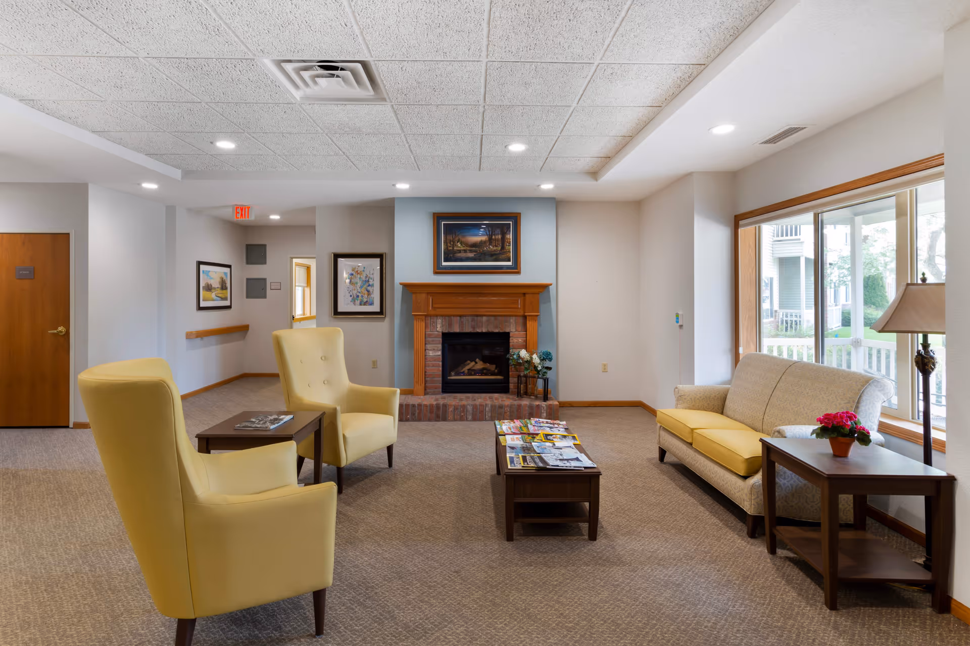 A cozy senior living facility common area with two yellow armchairs and a beige sofa arranged around a wooden coffee table covered with magazines. A brick fireplace with a wooden mantle and framed artwork above it is centered on the far wall. Large windows on the right side let in natural light, and there are side tables with a lamp and a potted plant. The room has carpeted floors, recessed ceiling lights, and neutral-colored walls.