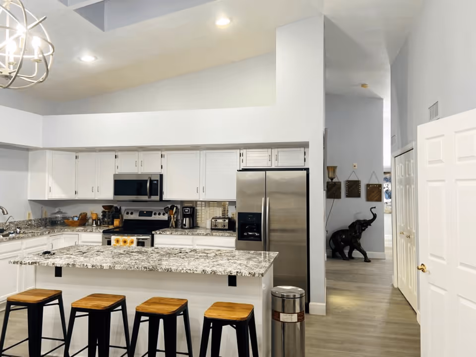 Bright modern kitchen with a large granite island, four bar stools, stainless steel appliances, and white cabinetry.