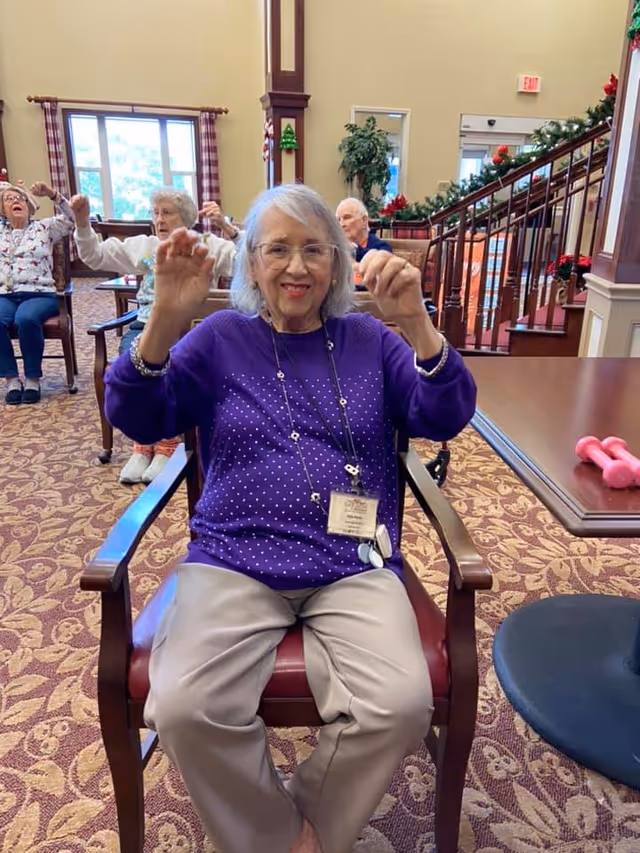 An elderly woman wearing a purple sweater and beige pants is seated in a chair in a common area, smiling and raising her hands as if exercising. Behind her, other elderly individuals are also seated and participating in the activity. The room has patterned carpet, large windows with checkered curtains, and a staircase decorated with garlands and red bows.