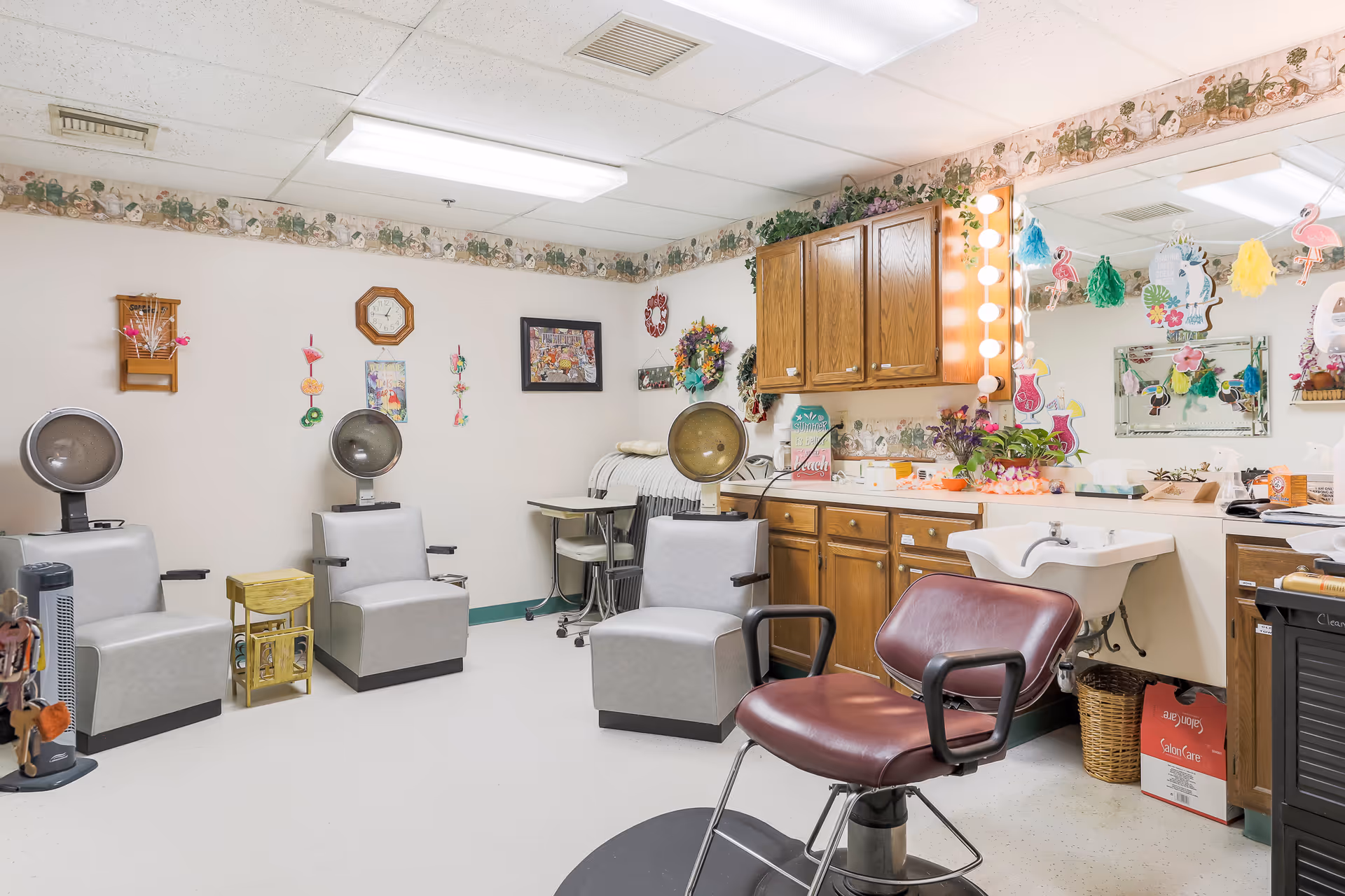 Interior of a senior living facility's hair salon area with three gray salon chairs, one maroon salon chair, a sink, wooden cabinets, a large mirror decorated with colorful hanging ornaments, and various hair care items on the counter.