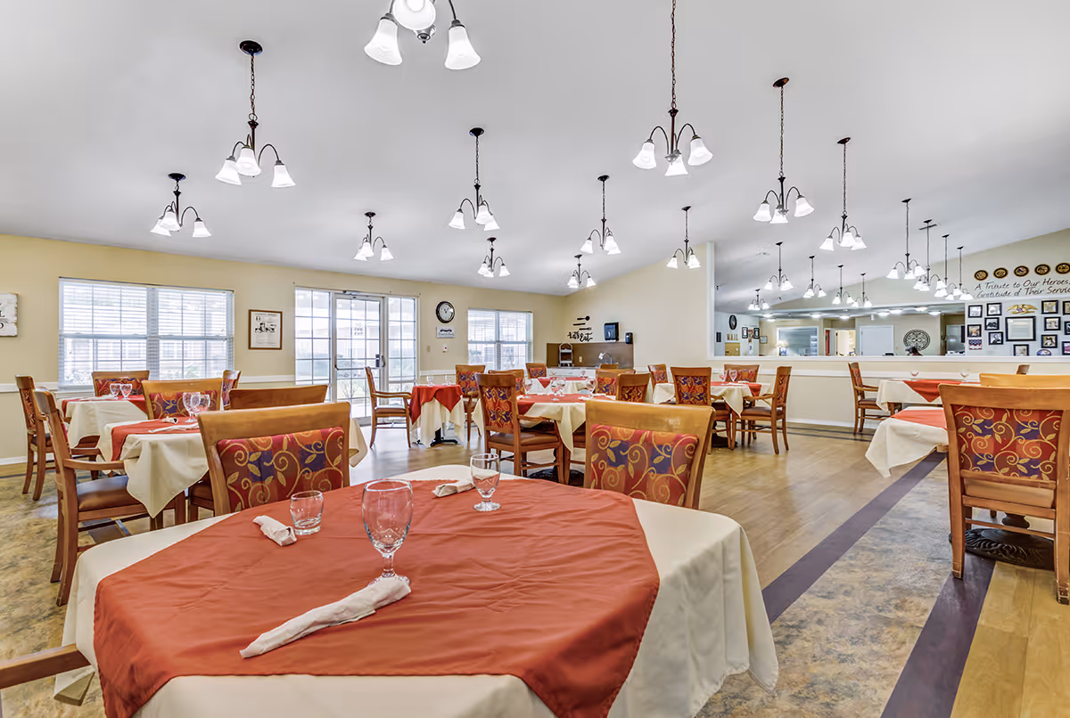 A spacious dining room in Meadowview Place with multiple tables covered in white and red tablecloths, each set with glasses and napkins. The room has wooden chairs with patterned cushions, large windows letting in natural light, and multiple ceiling light fixtures. A large mirror on one wall reflects the room, and there are framed pictures and a tribute on the far wall.