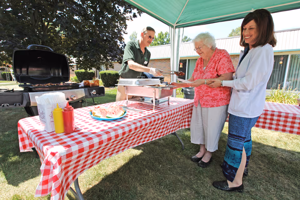An outdoor barbecue event at a senior living facility with a man serving food to an elderly woman holding a plate, while a smiling woman stands beside her. The table is covered with a red and white checkered tablecloth and has condiments, napkins, and a plate of watermelon slices. There is a grill and a canopy providing shade, with a building and greenery in the background.