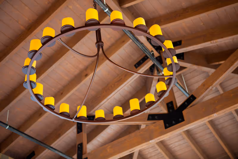 Circular chandelier with multiple yellow candle-like lights hanging from a wooden ceiling with exposed beams and metal supports.