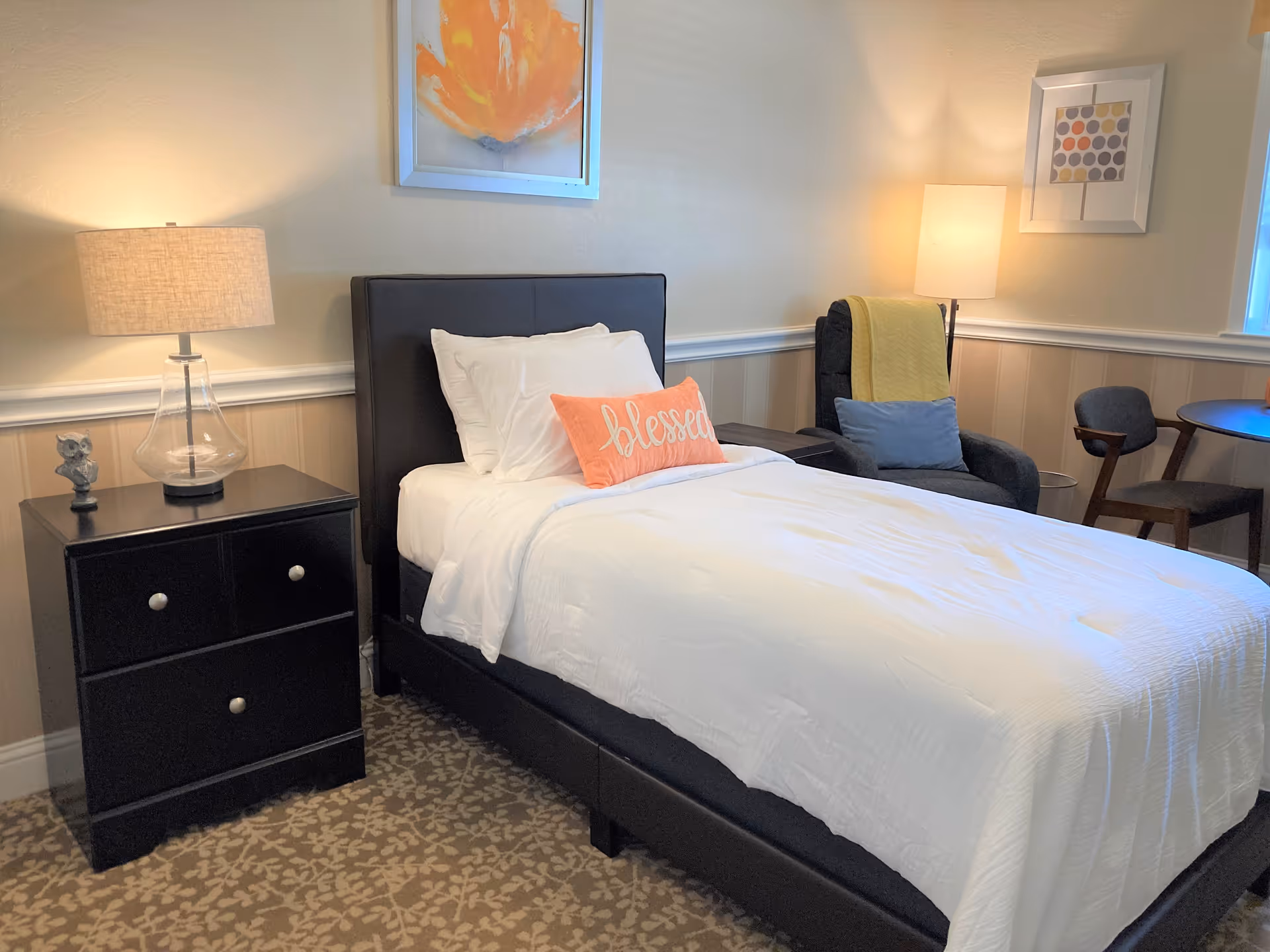 A neatly made single bed in a decorated bedroom with a nightstand and lamp, armchair, and small table.