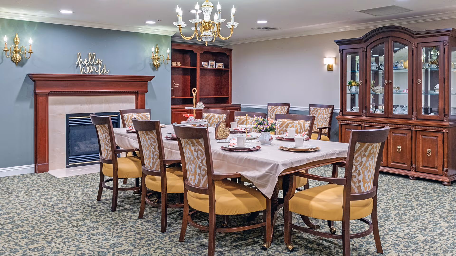 A formal dining room with a large rectangular table covered with a white tablecloth, set with plates, cups, and napkins. The room features eight wooden chairs with yellow cushioned seats and patterned backs. There is a wooden fireplace with a 'hello world' sign above it on a green accent wall, a wooden china cabinet displaying dishes, and a wooden bookshelf. The room is carpeted with a patterned design and illuminated by a chandelier and wall sconces.