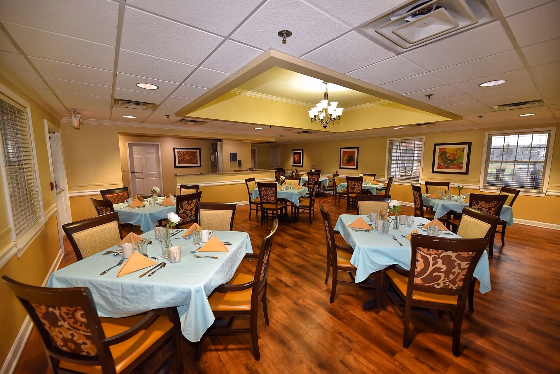 A dining room with multiple tables covered in light blue tablecloths, each set with orange folded napkins, cups, glasses, and silverware. The room has wooden floors, beige walls with framed artwork, and windows with blinds. A chandelier hangs from the ceiling, and the room is well-lit with recessed lighting.