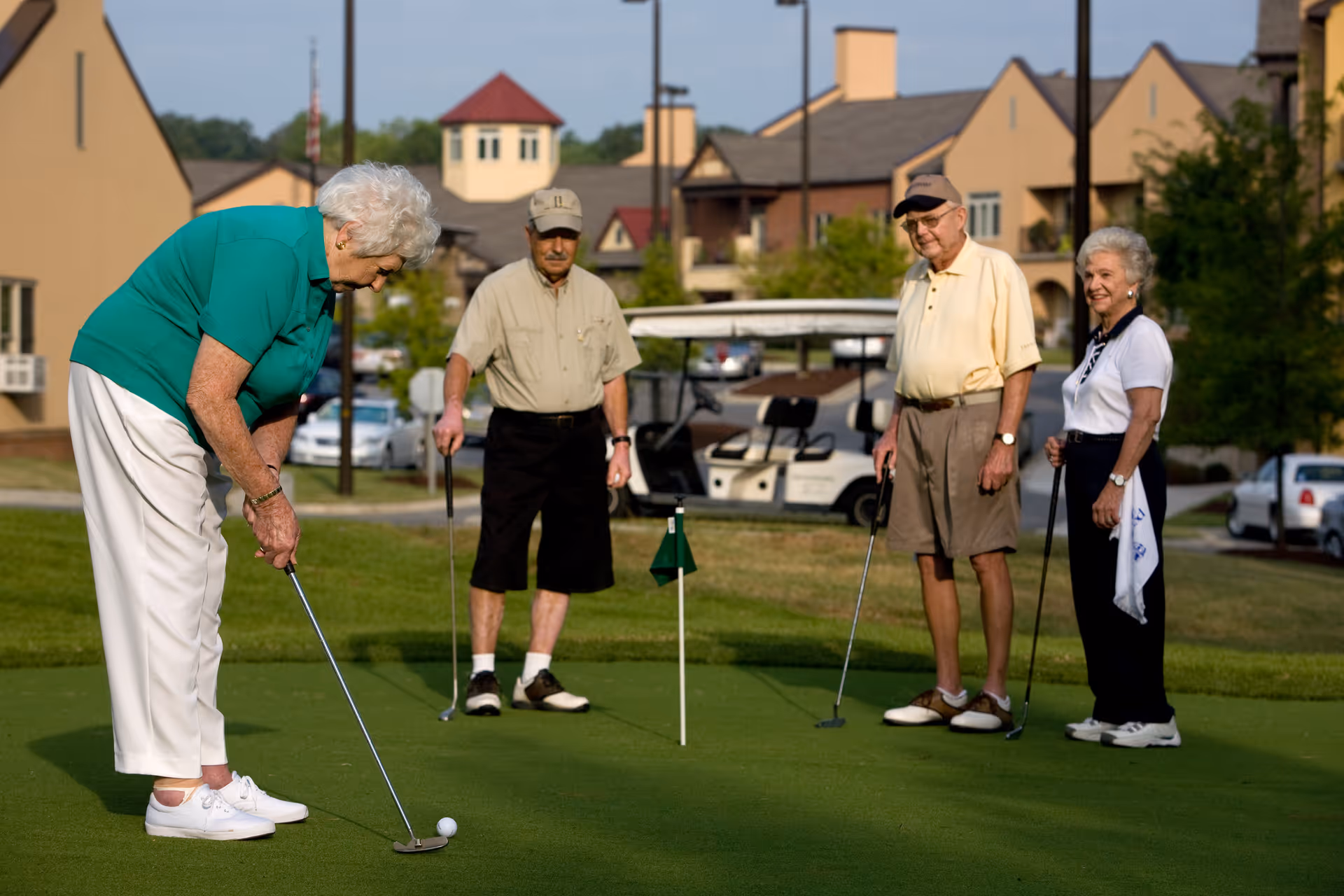 Four elderly people playing golf on a putting green. One woman in a green shirt and white pants is putting the ball, while two men and another woman stand nearby watching. There are buildings and a golf cart in the background.
