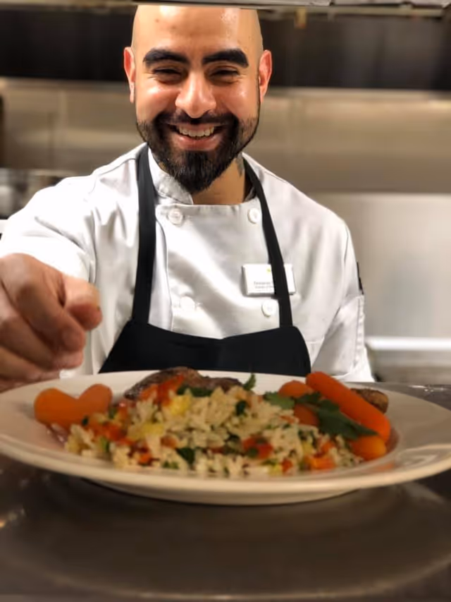A smiling chef wearing a white coat and black apron presents a plate of food with rice, vegetables, and carrots in a kitchen setting.