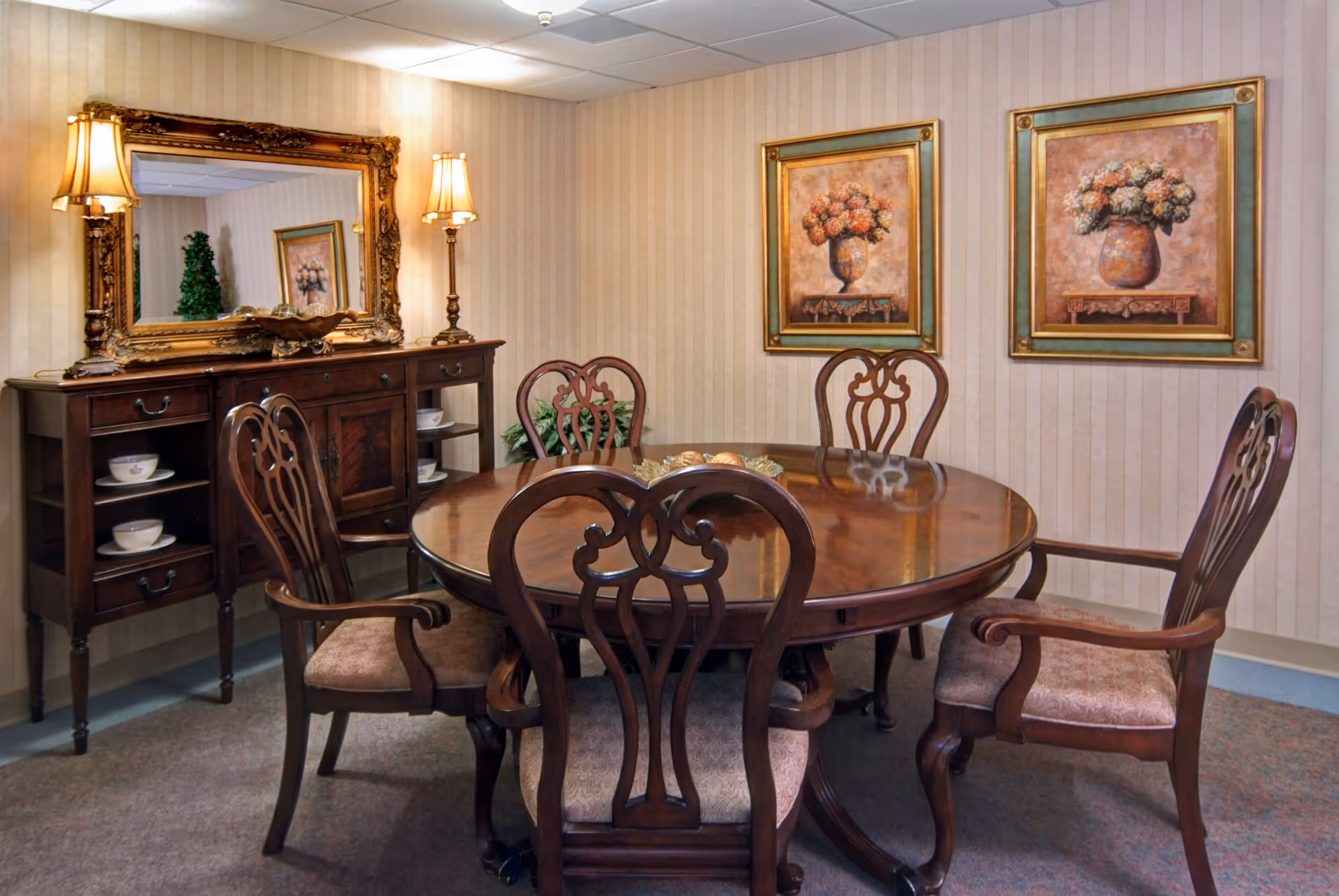 A formal dining room with a round polished wooden table surrounded by carved chairs, a sideboard with a mirror and lamps, and two framed floral paintings on the wall.