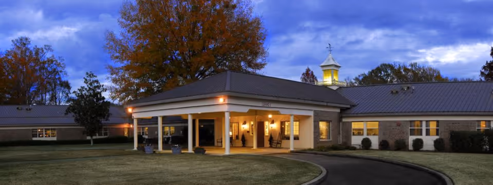 Front entrance of a single-story brick senior living building with a covered porte-cochere, cupola, lit windows and a curved driveway at dusk.