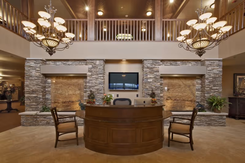 A spacious and elegant reception area with a curved wooden desk and two wooden chairs. Behind the desk are two stone water features and a mounted flat-screen TV. The area has a high ceiling with two ornate chandeliers and a balcony railing above.
