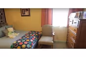 A small bedroom with a bed on the left side covered with a colorful crocheted blanket and a teddy bear sitting on the pillow. There is a wooden chair with armrests placed in front of a window with orange curtains. On the right side, there is a tall wooden dresser with some items on top.