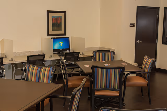 A room with several tables and chairs featuring striped upholstery. In the background, there is a computer workstation with a monitor displaying the Windows desktop screen, and privacy dividers between the workstations. A framed artwork hangs on the wall above the computer area, and a closed dark wooden door is visible on the right side.