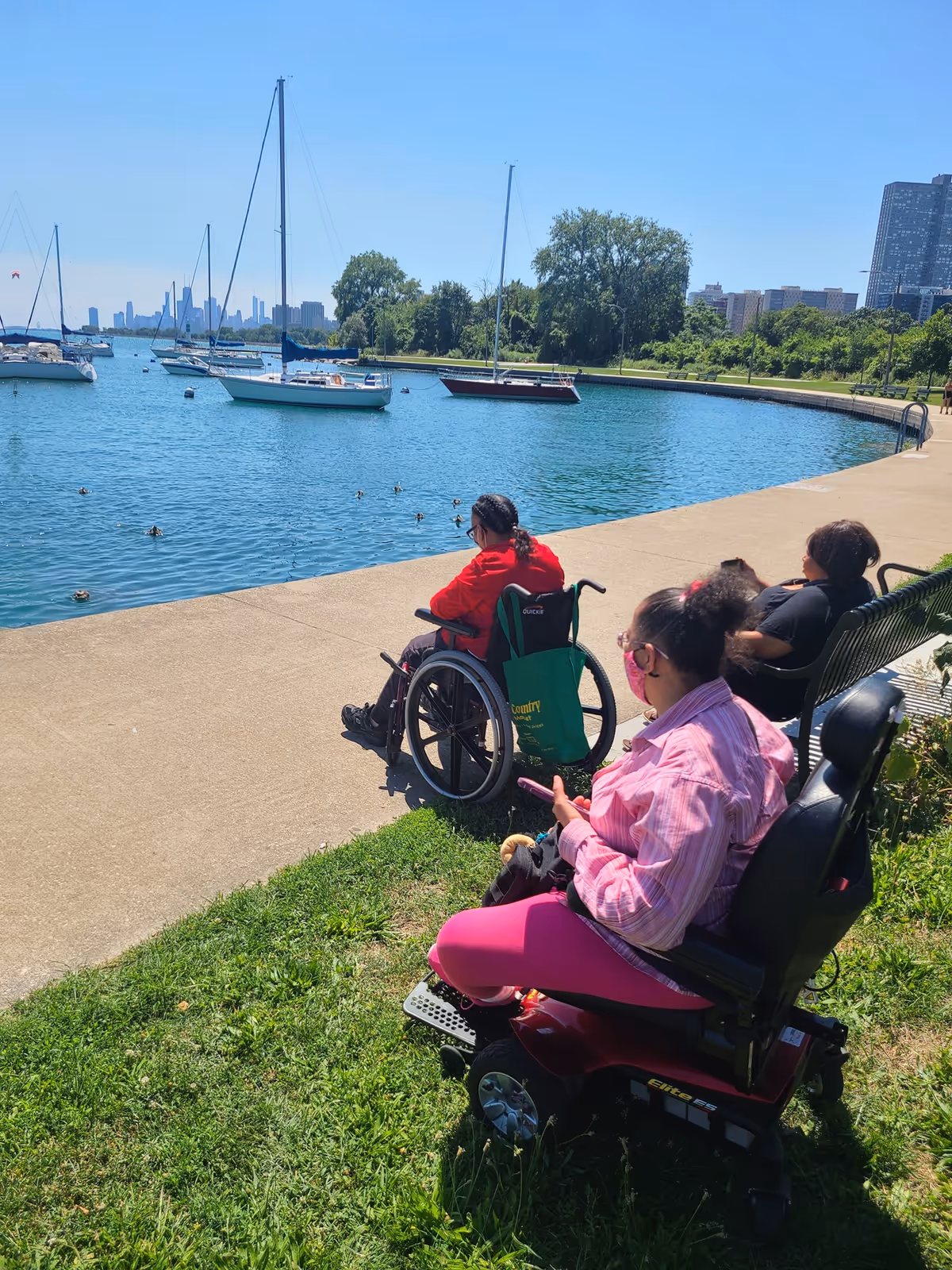 Three people near a waterfront on a sunny day. One person in a wheelchair wearing a red jacket is sitting on the paved path by the water. Another person in a pink jacket and pink pants is seated in a motorized wheelchair on the grass, holding a phone. A third person is sitting on a bench facing the water. Several sailboats are anchored in the water, and trees and buildings are visible in the background.