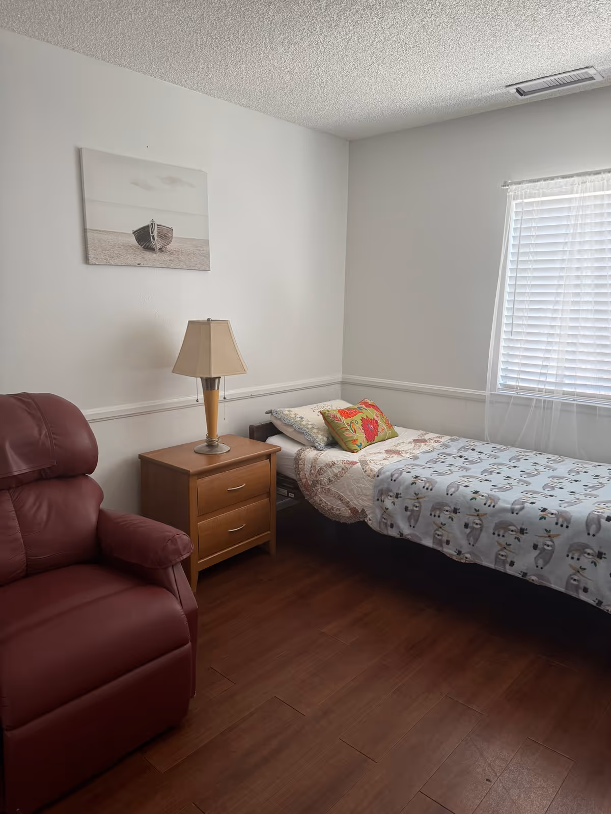 Small bedroom with a single bed next to a wooden nightstand and lamp, a red recliner, wall art, and a window with blinds.
