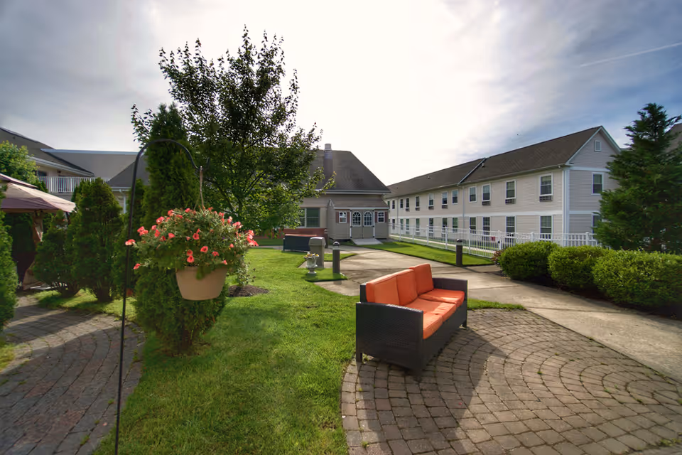 Outdoor garden area at Rose Garden Nursing and Rehabilitation Center featuring a paved circular seating area with an orange cushioned bench, green grass, hanging flower pot with pink flowers, trees, shrubs, and a two-story building in the background under a partly cloudy sky.