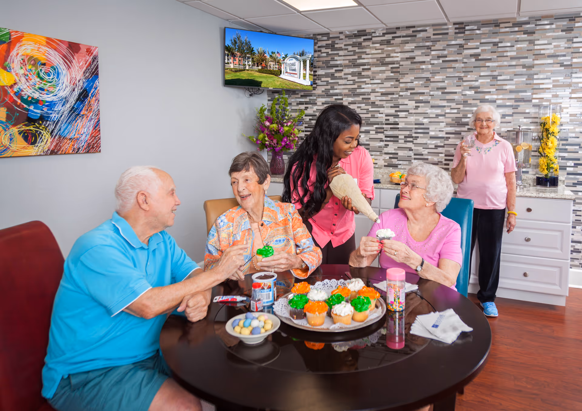 A group of elderly people and a caregiver decorating and enjoying cupcakes around a round table in a bright room with a tiled backsplash. The caregiver is piping frosting onto a cupcake held by an elderly woman in a pink shirt, while two other elderly people sit nearby holding decorated cupcakes. Another elderly woman stands in the background near a counter with a beverage dispenser and flowers. A colorful abstract painting hangs on the wall and a TV screen displays an outdoor scene.