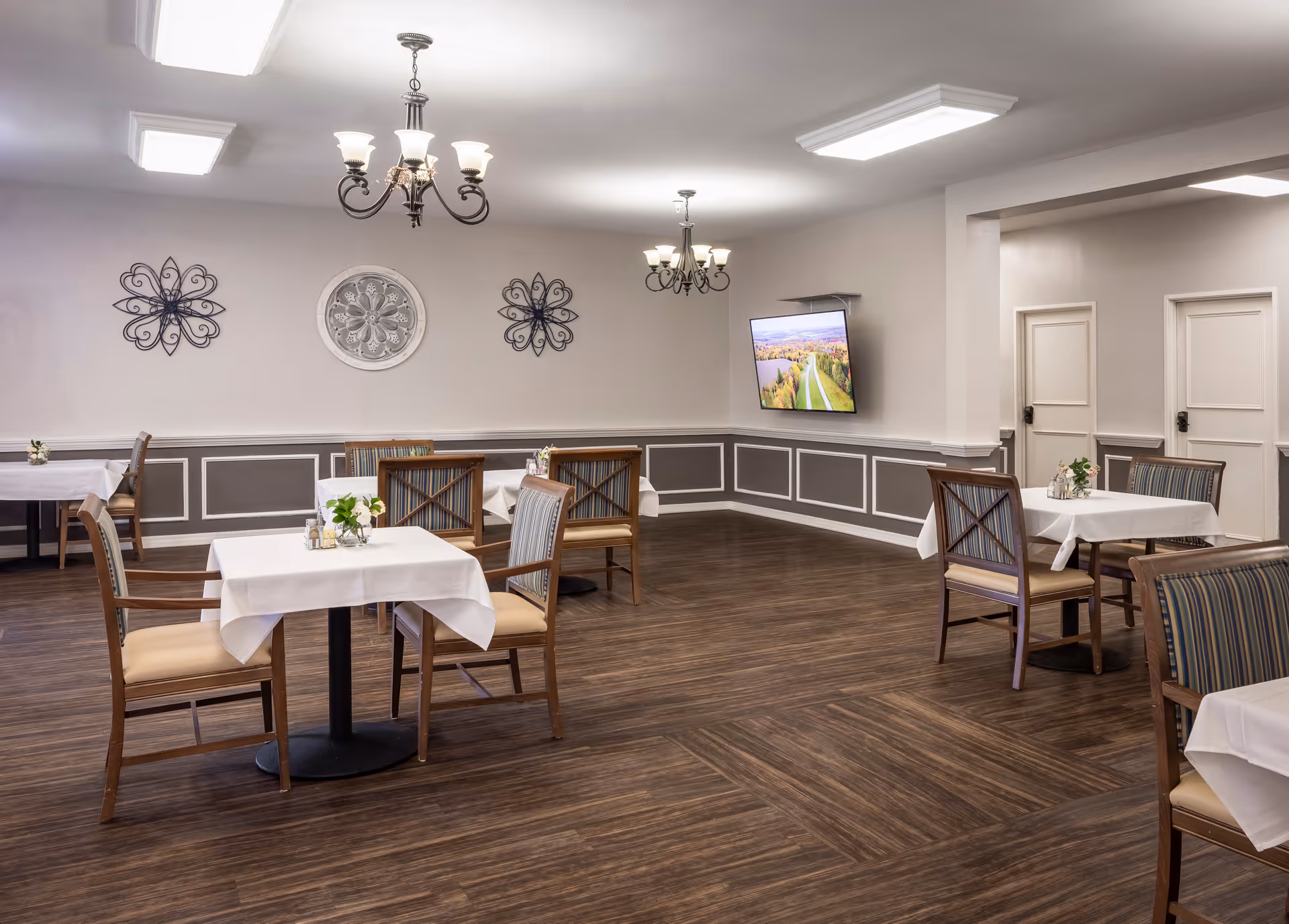 A dining room with several tables covered in white tablecloths, each surrounded by wooden chairs with cushioned seats. The room has wood flooring, gray and white paneled walls, decorative wall art, two chandeliers, and a wall-mounted flat-screen TV displaying a scenic landscape.