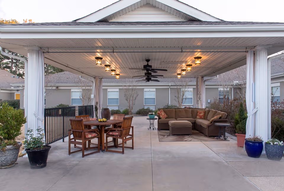 Outdoor covered patio area with ceiling fans and hanging lights. There is a round wooden table with four chairs on the left side and a brown sectional sofa with cushions on the right side, placed on a rug. Several potted plants are arranged around the patio, and a building with windows is visible in the background.