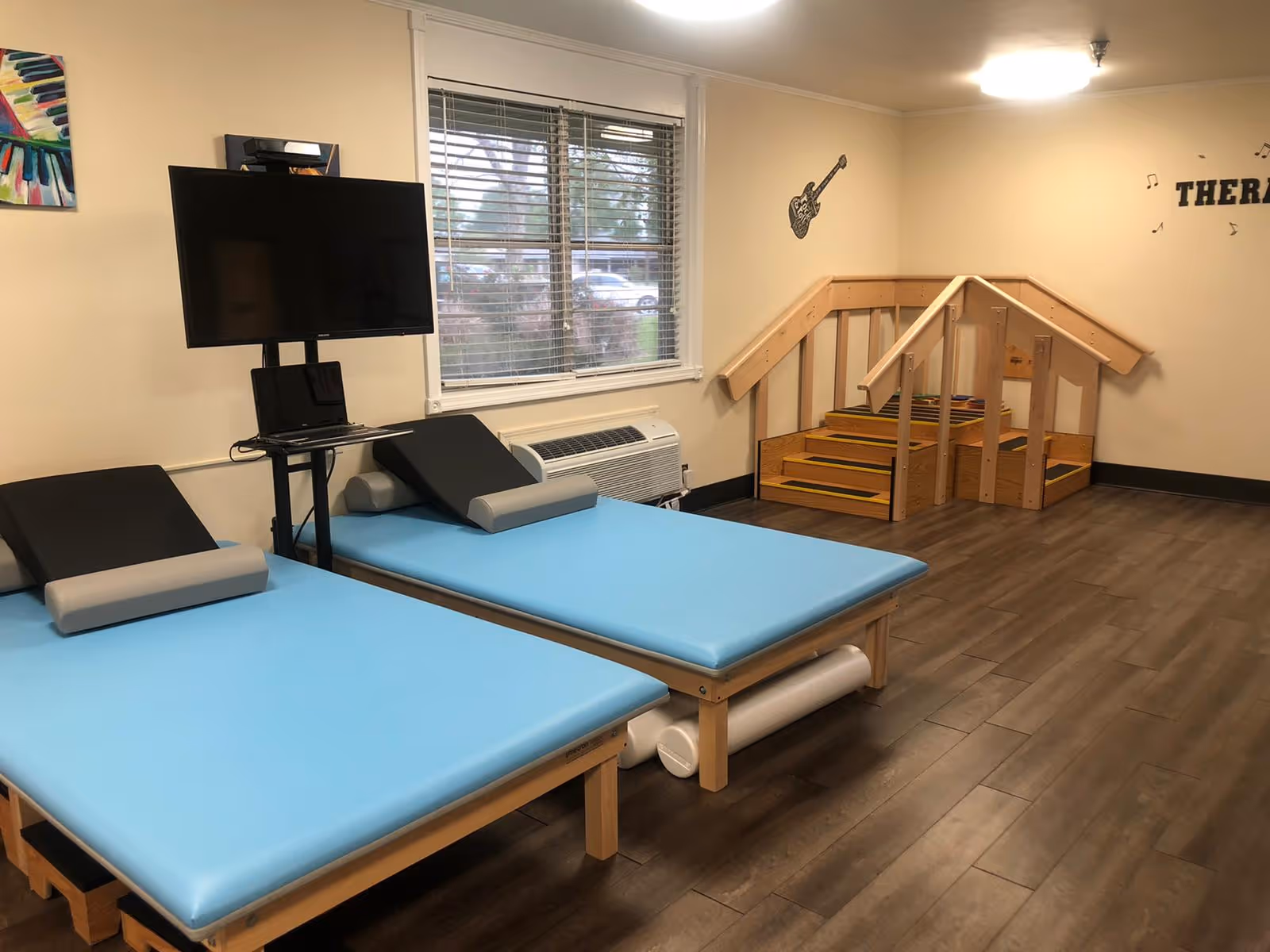 A therapy room with two blue padded therapy tables, a TV mounted on a stand, a window with blinds, and a small wooden staircase with handrails. The walls are beige with a guitar decoration and the word 'THERAPY' partially visible.