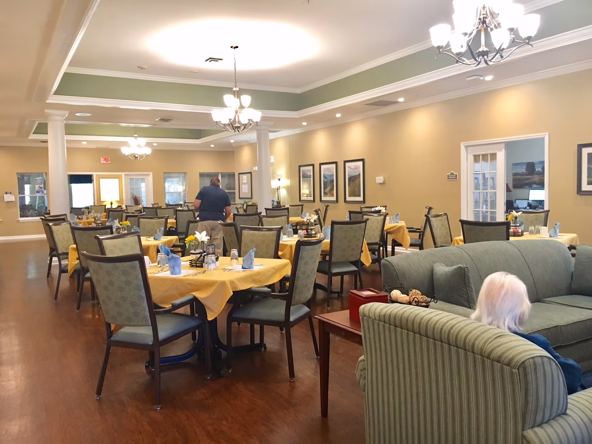 A spacious dining room in a senior living facility with multiple tables covered in yellow tablecloths, each set with blue napkins, glasses, and small flower arrangements. A staff member is seen attending to one of the tables. In the foreground, an elderly person with white hair is seated on a green striped couch. The room has warm beige walls, framed landscape pictures, and elegant chandeliers hanging from a tray ceiling with green accents.