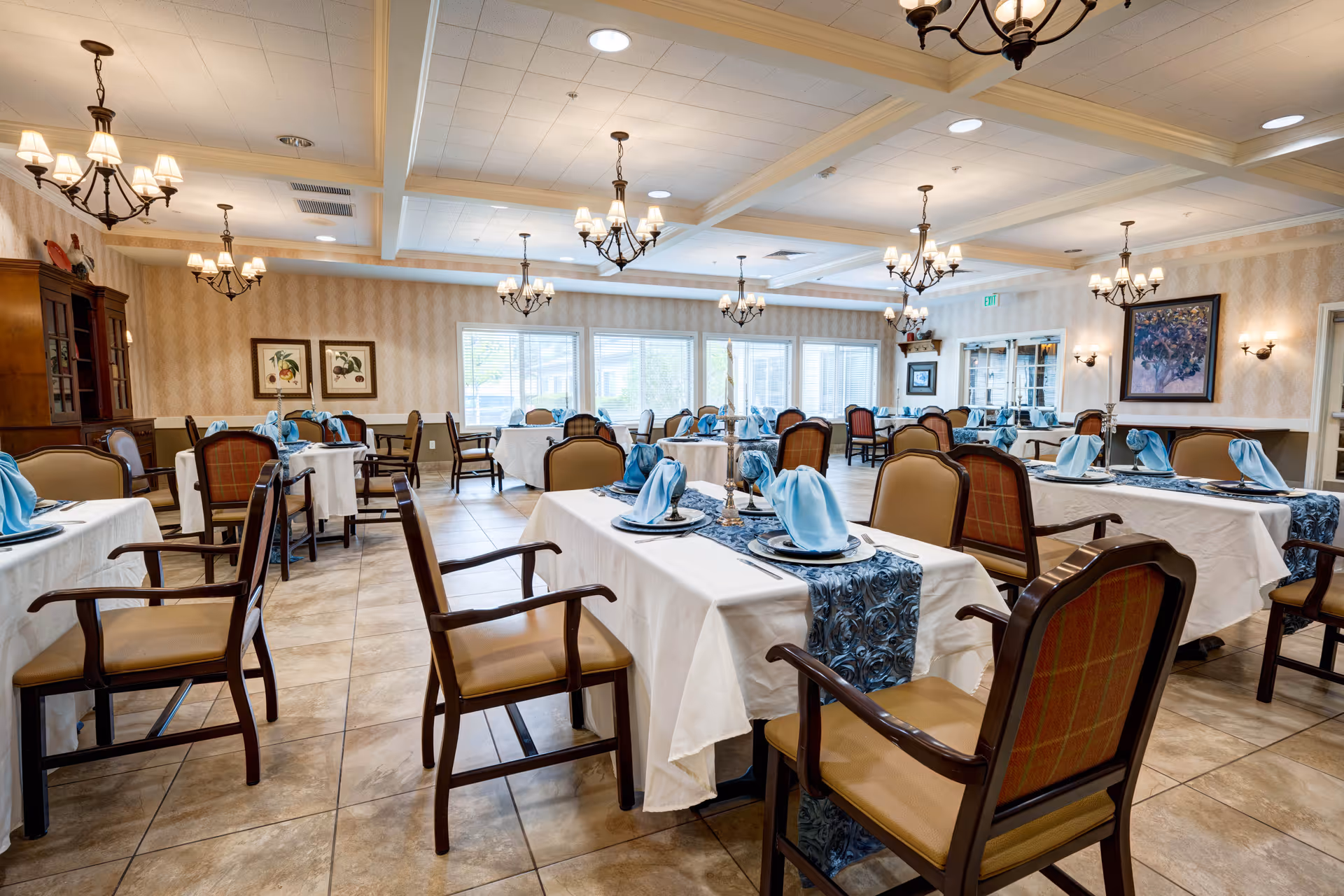 A spacious dining room with multiple tables covered in white tablecloths and blue patterned runners. Each table is set with plates and blue folded napkins. The room features wooden chairs with cushioned seats and backs, chandeliers hanging from the ceiling, framed artwork on the walls, and large windows letting in natural light.