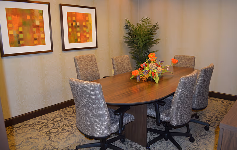 A small conference or meeting room with a wooden oval table surrounded by six cushioned office chairs on wheels. A floral arrangement with orange flowers is placed in the center of the table. The room has patterned carpet flooring, beige walls with two framed abstract artworks, and a tall green potted plant in the corner.