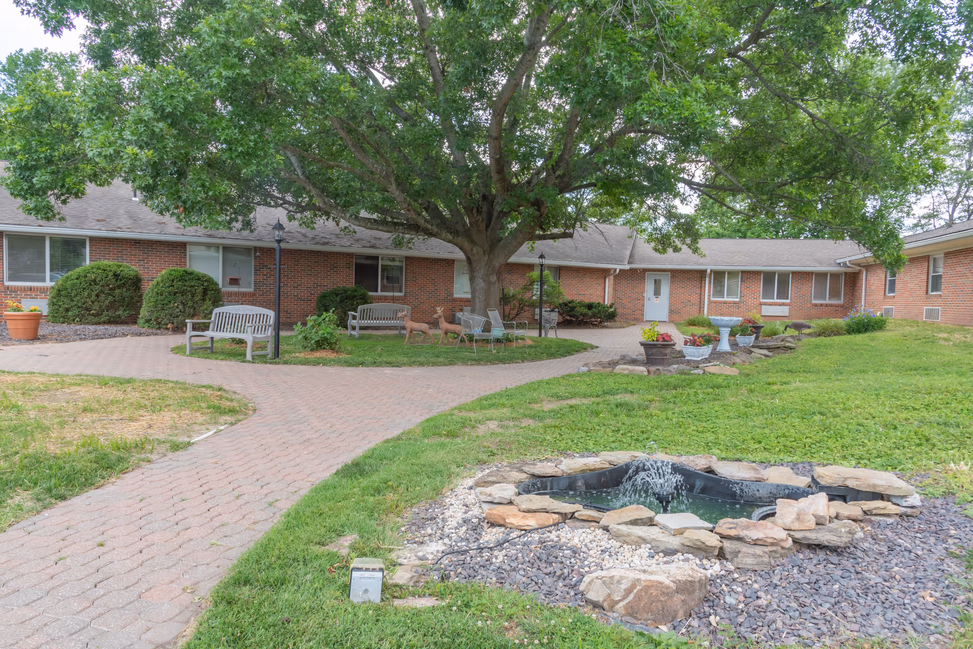 Outdoor courtyard area of a senior living facility with a large tree in the center, surrounded by benches, chairs, and decorative deer statues. There is a small stone-bordered water fountain in the foreground and brick buildings with windows and doors in the background.