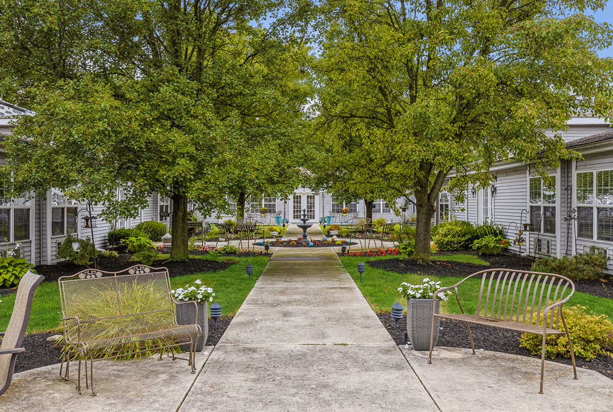 A peaceful outdoor courtyard at DeWolfe Place featuring a concrete walkway lined with small garden lights, green grass, flower beds, and several trees. There are metal benches and chairs arranged along the walkway, with a decorative fountain and additional seating visible in the background. The courtyard is surrounded by a building with large windows.