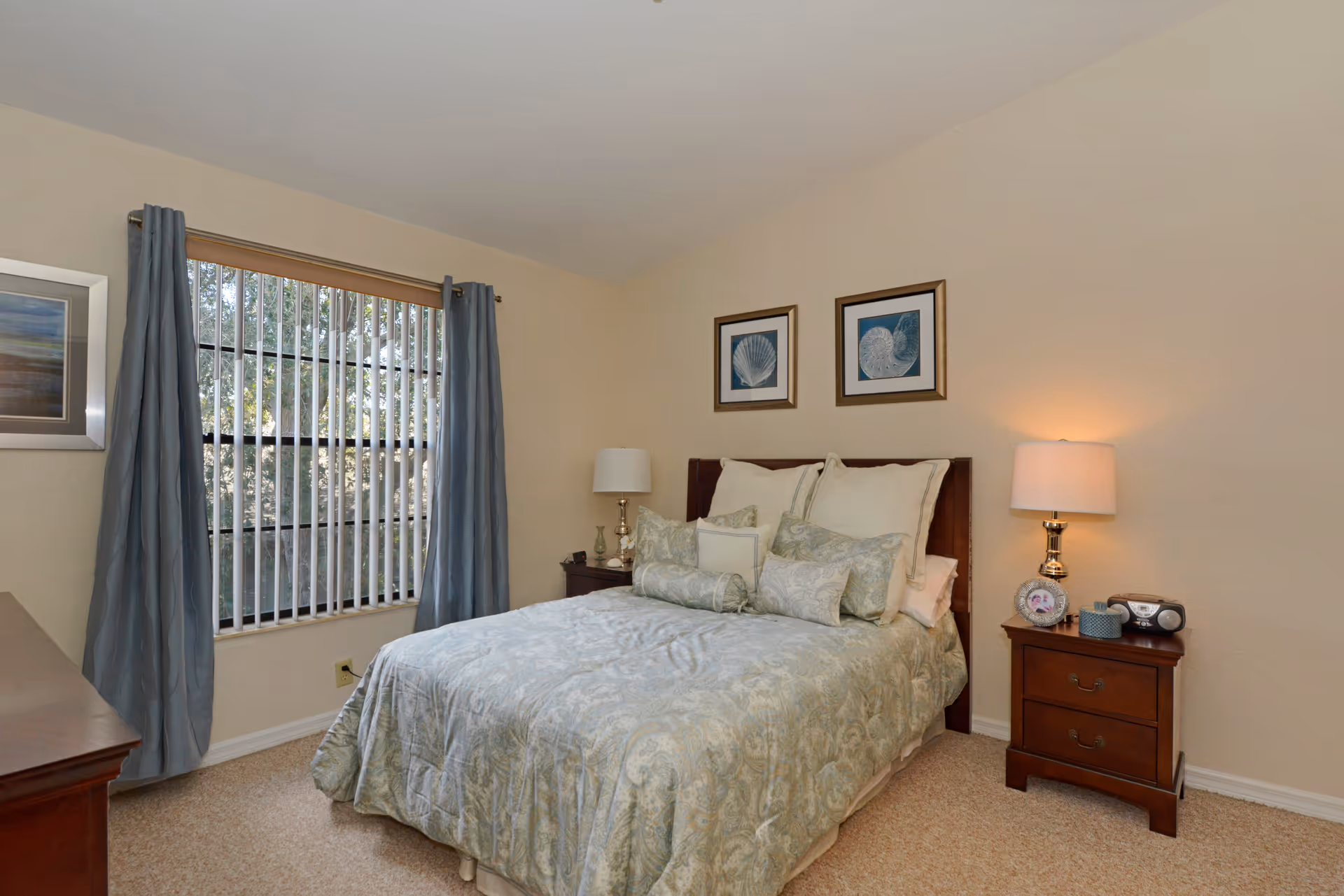 Neatly made bed with patterned bedding between two nightstands and lamps in a softly lit bedroom with a window with vertical blinds and blue curtains.