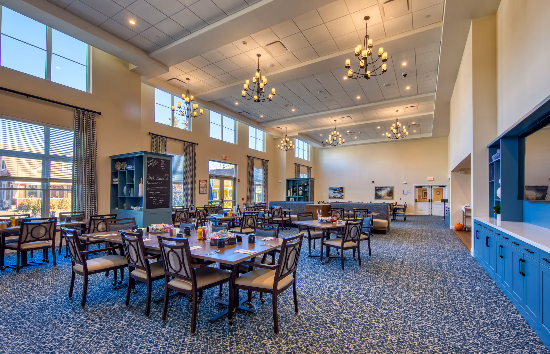 A spacious dining room with multiple tables and chairs arranged neatly. The room has large windows letting in natural light, blue cabinetry along one wall, and several chandeliers hanging from a high ceiling. The carpet has a patterned design, and there are shelves with decorative items and a chalkboard menu.