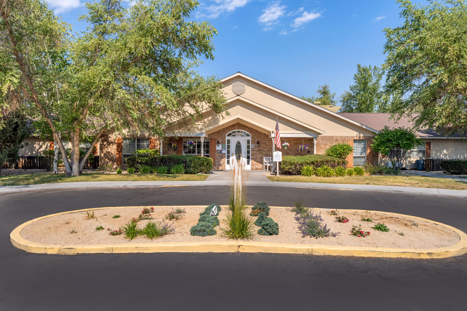 Front exterior view of a single-story senior living facility building with a peaked roof, beige and brick facade, and an American flag near the entrance. The building is surrounded by green trees and shrubs, with a landscaped island in the circular driveway in front.
