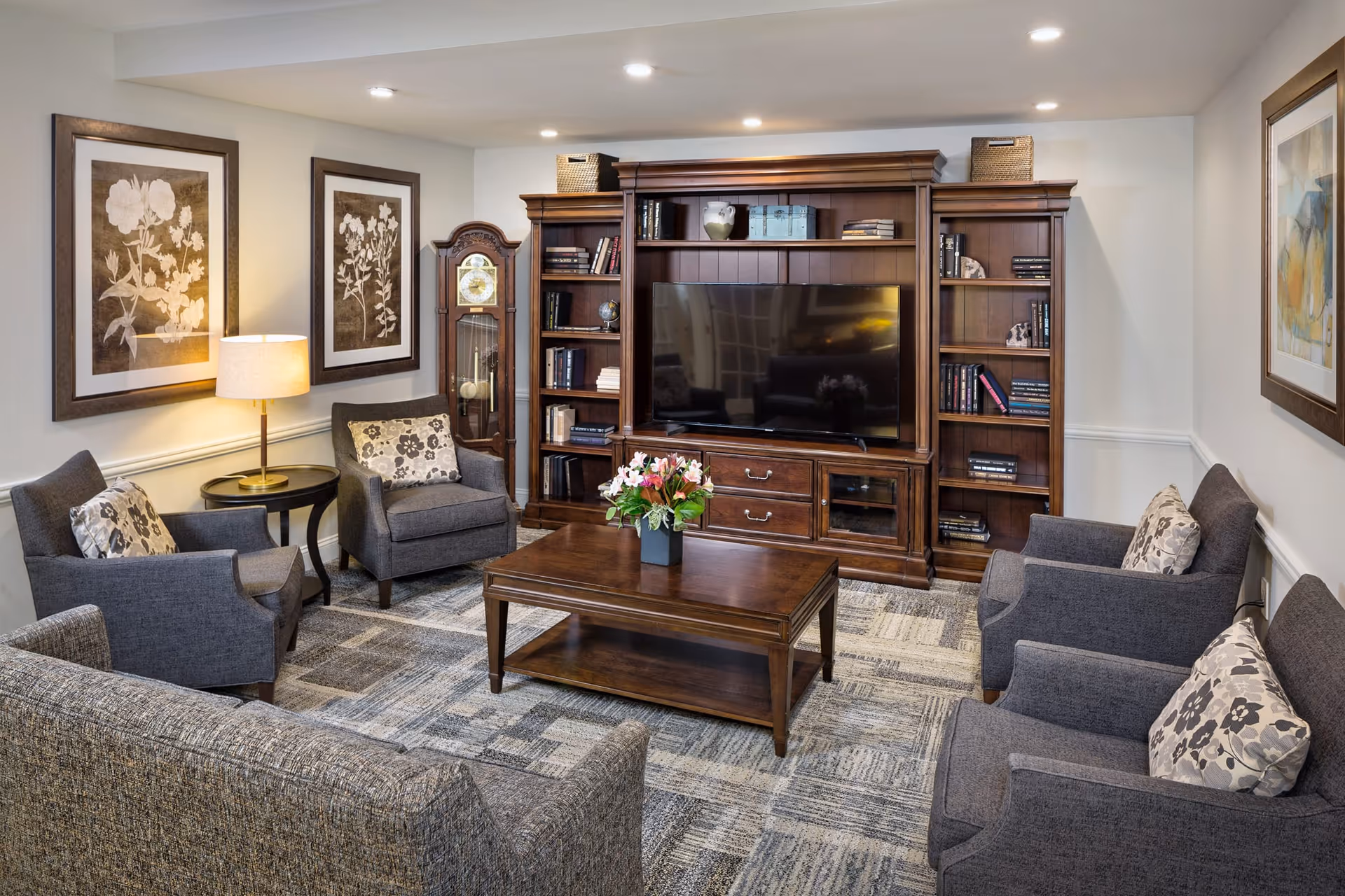 A cozy living room with a large wooden entertainment center holding a flat-screen TV and books. The room has four gray armchairs and a gray sofa with floral patterned cushions arranged around a wooden coffee table with a flower arrangement. The walls are decorated with framed artwork and a grandfather clock is placed in the corner. A table lamp provides warm lighting.