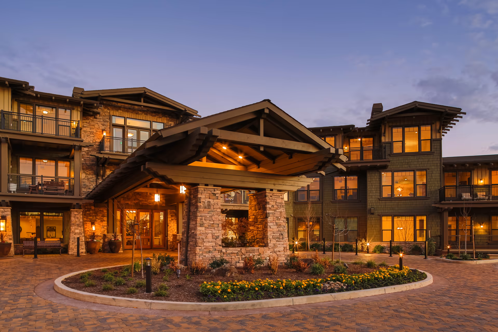 Exterior view of a senior living facility at dusk with warm lights glowing from the windows. The building features stone pillars and wooden architectural details with a covered entrance and landscaped flower beds in front.
