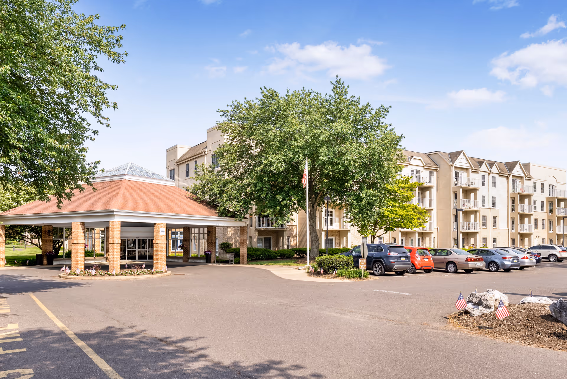 Front entrance of a multi-story senior living building with a covered porte-cochère, parked cars, and trees.